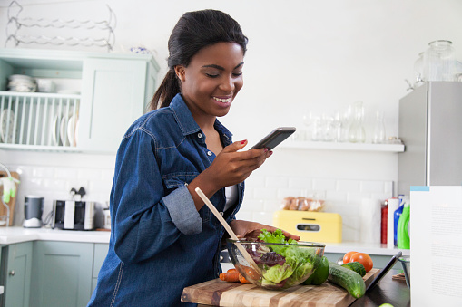 African female looking at her cellphone with the biggest smile after reading a text message she received while cooking lunch in the kitchen. Cape Town, Western Cape, South Africa.