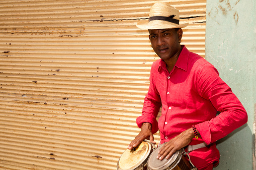 A well known professional Cuban conga drum player percussionist and the afro-cuban Santeria priest in red clothes and Panama hat standing against a closed yellow store shutter, playing bongo drums. A lot of copyspace. Old Havana, Cuba.
