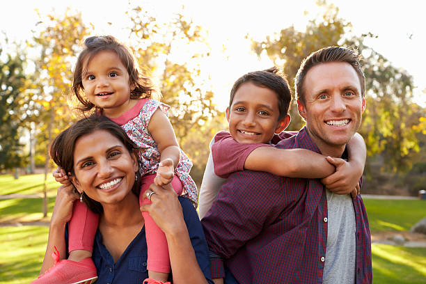 genitori di razza mista portano i loro figli piggyback in un parco - multi ethnic family foto e immagini stock