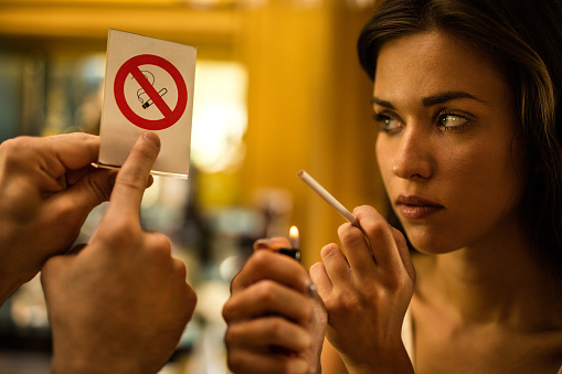 Young woman is about to light a cigarette while  hand of a man pointing at no smoking sign.