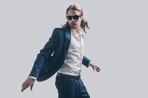 Handsome young man in full suit and sunglasses moving in front of grey background