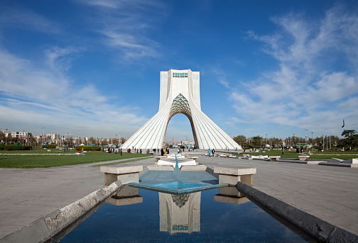 Azadi monument and its reflection on waterways in Azadi square of Tehran, against blue sky and white clouds.