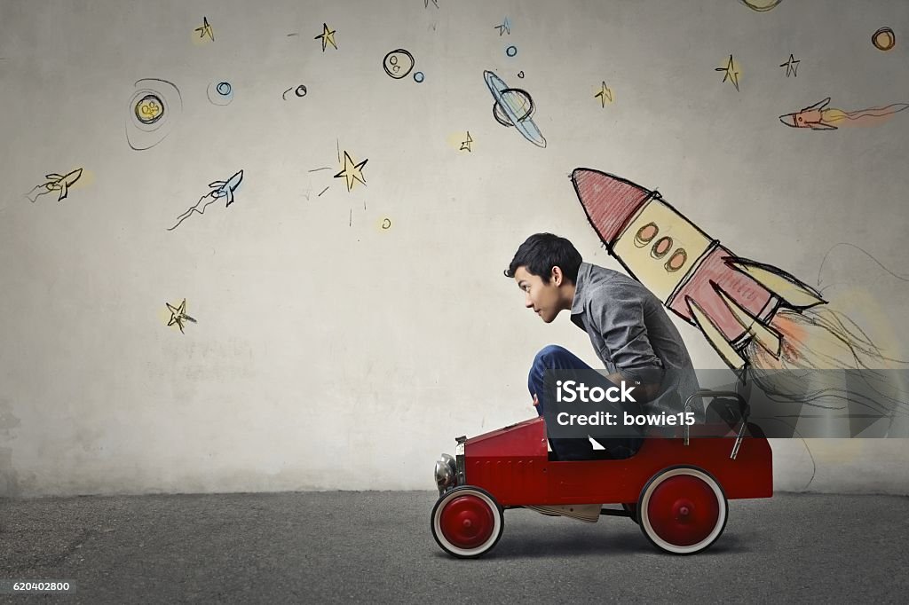To infinity and beyond The portrait of a young Asian guy in a red toy car in front of a gray wall with spaceship and planets. Child Stock Photo