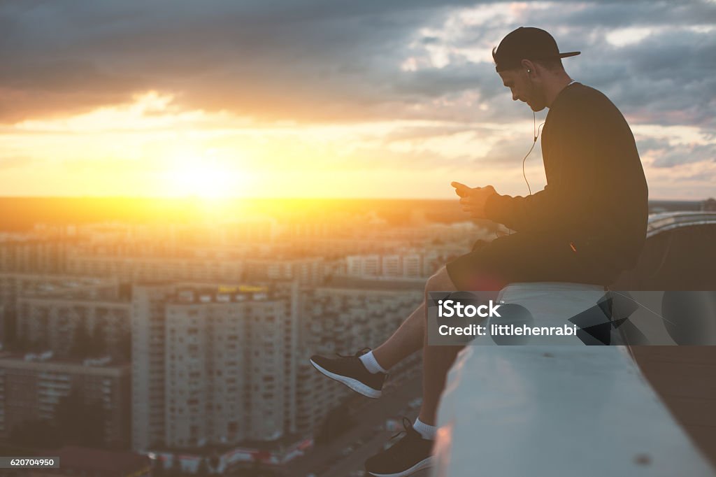 Hombre sentado en el borde del techo con teléfono inteligente - Foto de stock de Ciudad libre de derechos