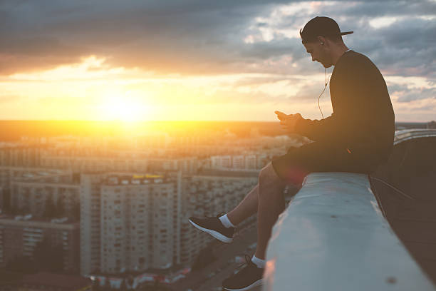 man sitting on the edge of the roof with smartphone - extreme sports audio imagens e fotografias de stock
