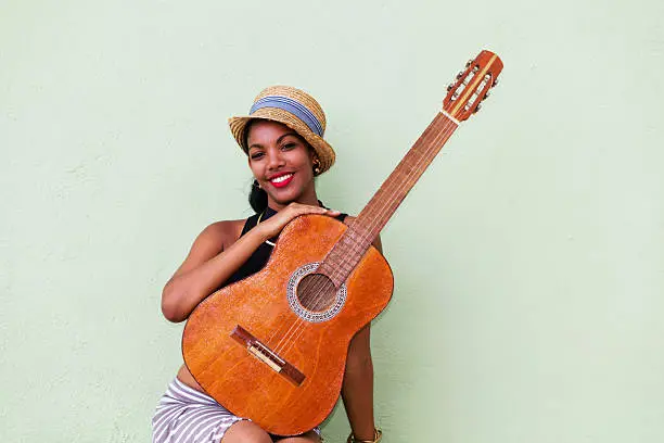 A portrait of a beautiful, young, smiling Cuban woman in Panama hat with a guitar in front of a green wall. Havana, Cuba, 50 megapixel image, copyspace.