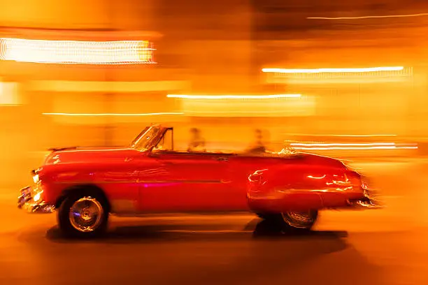Motion blurred vintage red American convertible car with unrecognizable people, speeding at night down the street in Havana, Cuba