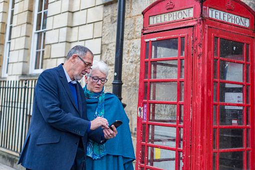 Senior adult couple using a mobile telephone outside a phone booth in Oxford, England in the UK