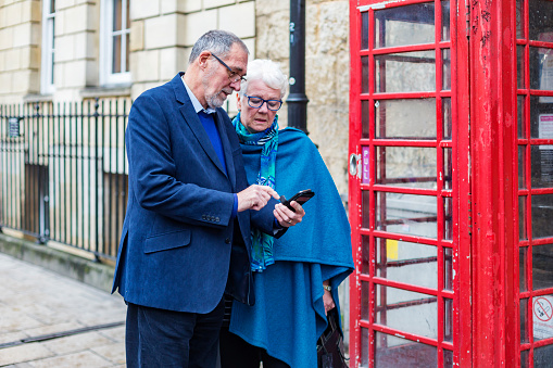Senior adult couple using a mobile telephone outside a phone booth in Oxford, England in the UK