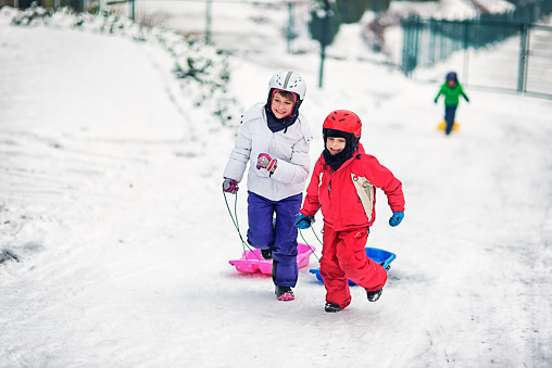 Three kids running up the hill with toboggans in a small town. The kids are wearing helmets.