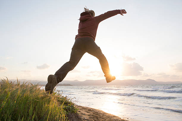 istock Young man leaps from beach dune, towards sea, sun 620969127