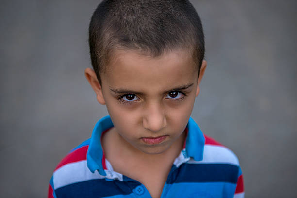 High angle view portrait of little boy standing in front of a blurred black background and looking at camera with disgusted facial expression. Horizontal composition. Image taken with Nikon D800 and developed from RAW format.