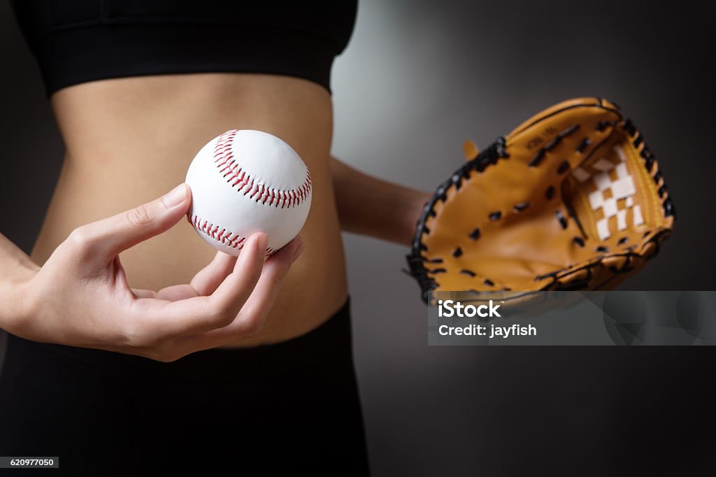 baseball glove and ball Close up studio shot of a fitness womans abdomen.  Fitness model holding a baseball glove and a white baseball. low key lighting shot on a grey background. Adult Stock Photo