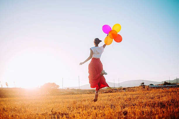 young woman joyfully jumping in the field holding colorful balloons - kvinna ballonger bildbanksfoton och bilder