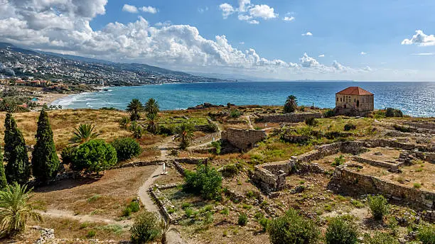 View over the roman ruins of the temple de Baccus, the little temple, in Byblos in Lebanon in the late afternoon. The sun reflects on the turquoise sea at the horizon providing, in the vivid summer light, a dramatic lighting and putting in evidence the shadows of the scenery of one of the most important archaeological areas of the mediterranean.