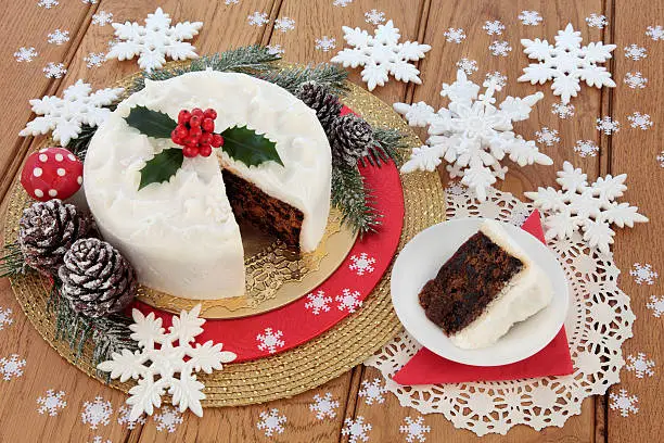 Traditional christmas cake and slice with holly, snow covered winter greenery with flly agaric mushroom bauble and pine cones with white snowflake decorations over oak background.