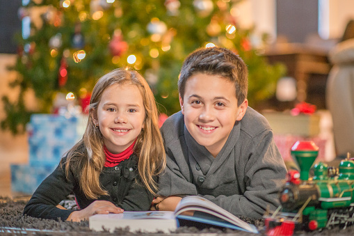 A brother and sister are reading a book by the Christmas tree. They are smiling and looking at the camera.
