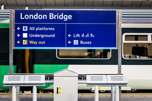 Horizontal colour image of a blue sign on the platform at London Bridge railway station in central London, UK. The sign has arrows pointing towards the other platforms, underground trains, the way out, buses and elevators. In the background a train is stationary, waiting for commuters to alight. 