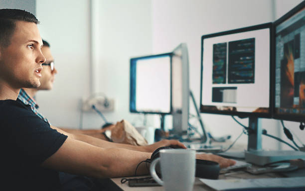 Software developers. Side view of two software developers sitting in front of  computers.They are looking at computer codes on the screen and thinking of how to do their task. man and machine stock pictures, royalty-free photos & images