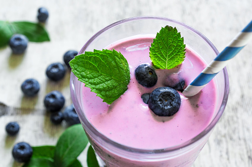 blueberry smoothie yogurt with fresh mint, paper straw and ripe berries on white wooden background