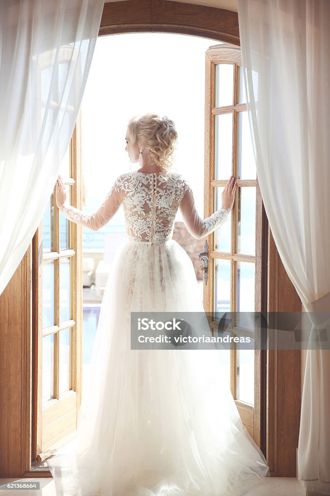 Young bride in gorgeous wedding dress with voluminous skirt Young bride in gorgeous wedding dress with voluminous skirt looking at window, indoors. Blonde young woman. Prom Dress Stock Photo