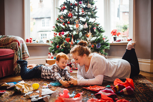 A young mother and her son are celebrating Christmas morning together, they are both lying on the floor and playing with newly opened toys together.