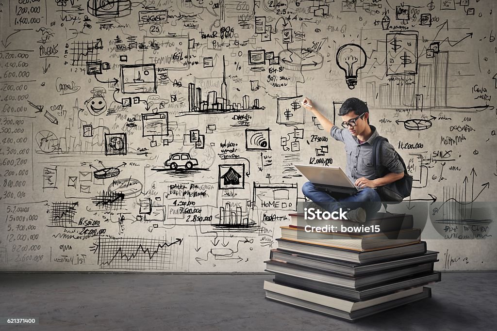 The more you know the more you can do Young Asian boy with nerdy black glasses sitting on a book hill, drawing his ideas creatively on the wall behind him while looking at his computer. Learning Stock Photo
