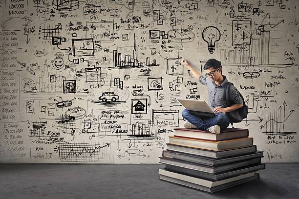 The more you know the more you can do Young Asian boy with nerdy black glasses sitting on a book hill, drawing his ideas creatively on the wall behind him while looking at his computer. nerd stock pictures, royalty-free photos & images