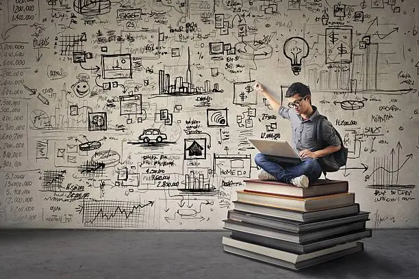 Young Asian boy with nerdy black glasses sitting on a book hill, drawing his ideas creatively on the wall behind him while looking at his computer.