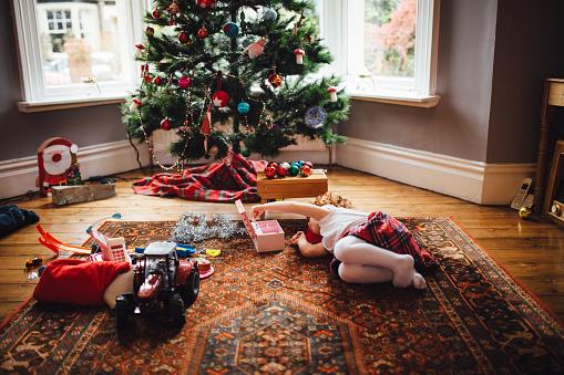 Christmas morning and a little girl is lying on the floor in front of the Christmas tree. The little redheaded girl is lying on her side playing with her new toys . A Christmas tree can be seen in the background and toys scattered around the floor.