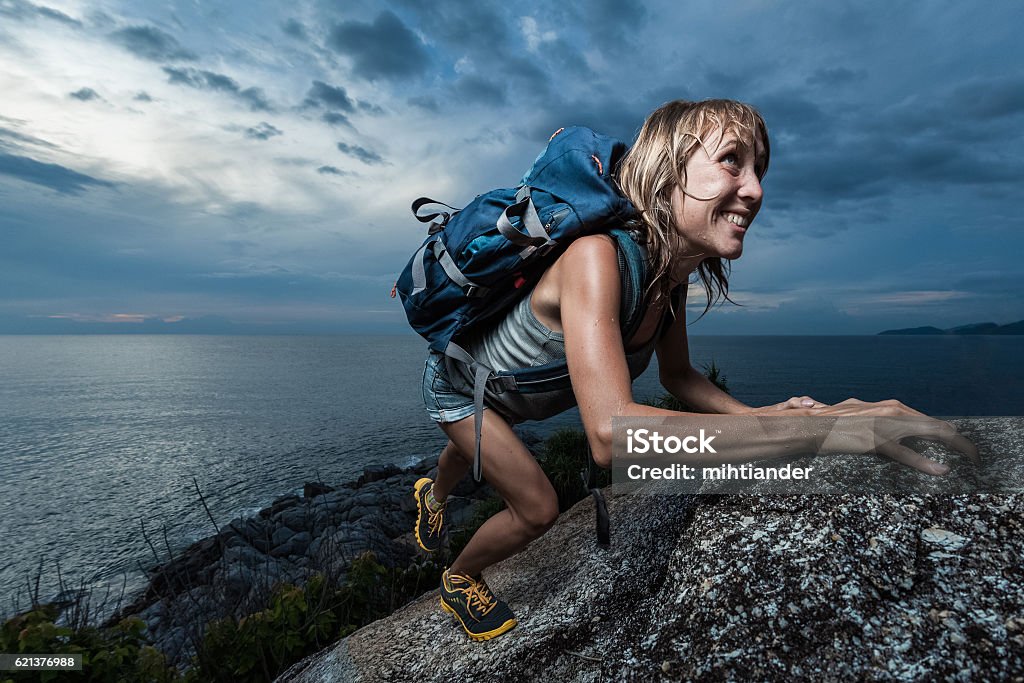 Lady on the rock Hiker with backpack climbing natural rocky wall on a dark cloudy background. There are water drops on the skin Sweat Stock Photo