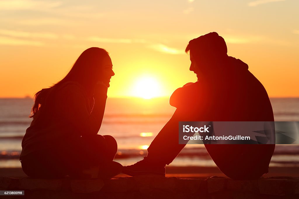 Friends or couple of teens facing at sunset Side view of full body of two friends or couple silhouette of teens facing at sunset on the beach with the sun in the middle Love At First Sight Stock Photo