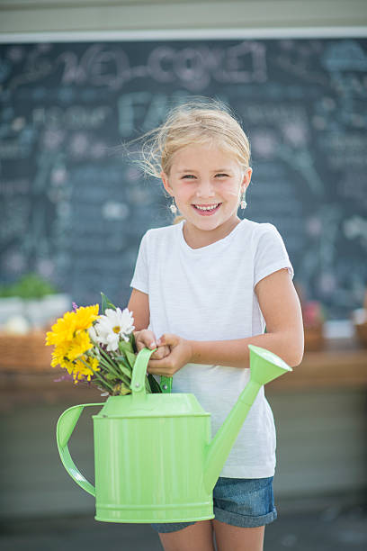 A little girl is selling daisies at the farmer's market. She is holding her flowers in a garden pale and is smiling while looking at the camera.