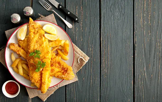 Two pieces of battered fish on a plate with chips on a wooden table with space