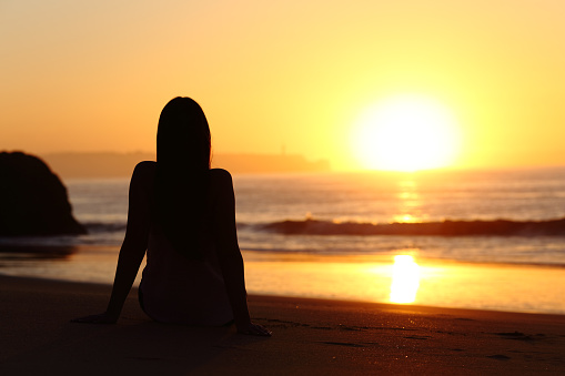 Back view of a woman silhouette sitting on the sand of a beach watching sun at sunrise with the horizon and ocean in the background