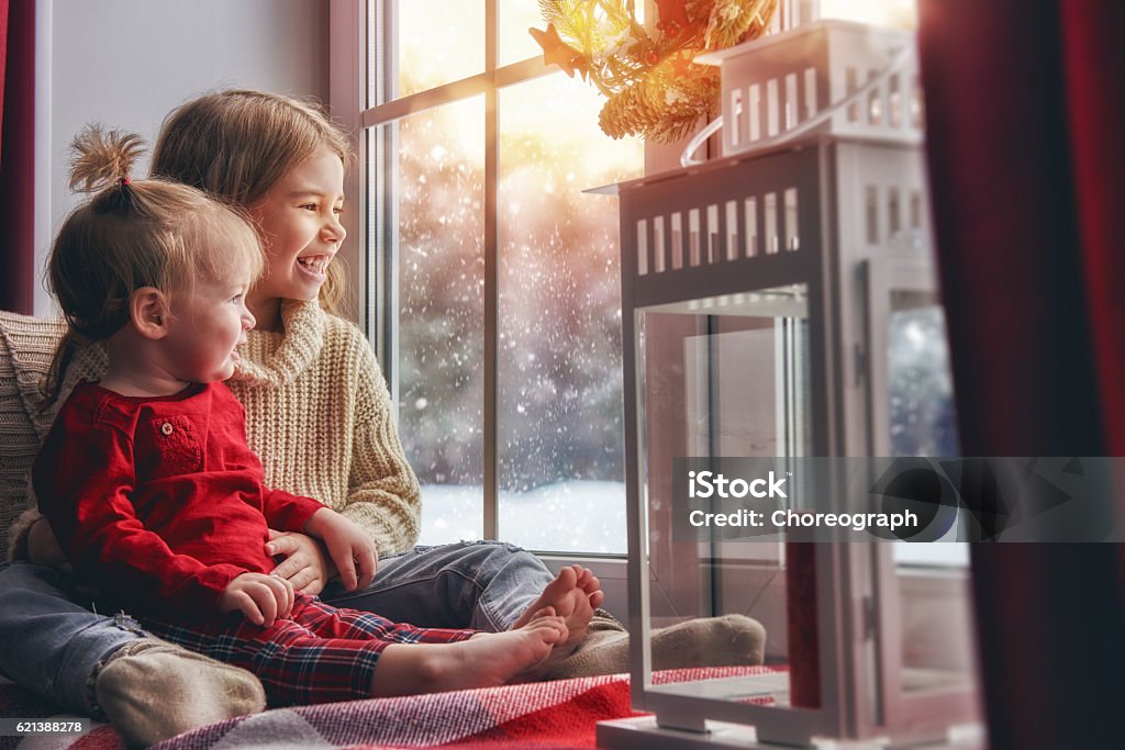 Kids enjoy the snowfall Merry Christmas and happy holidays! Cute little children girls sitting by the window and looking at the winter forest. Room decorated on Christmas. Kids enjoy the snowfall. Heat - Temperature Stock Photo