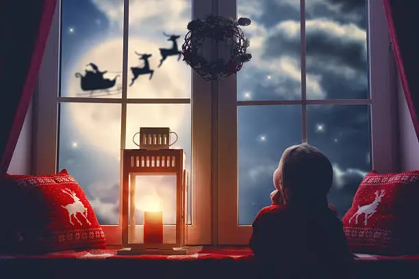 Merry Christmas and happy holidays! Cute little child girl sitting by window and looking at Santa Claus flying in his sleigh against moon sky. Room decorated on Christmas. Kid enjoy the holiday.