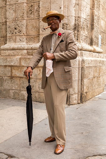 Elegant, elderly Cuban gentleman in a suit, straw boater hat, with a cigar and umbrella, standing in front of a wall in Havana, Cuba