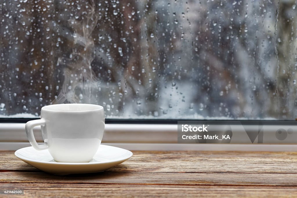 White cup on wooden windowsill White steaming cup of hot tea or coffee on vintage wooden windowsill or table against window with raindrops on blurred background. Shallow focus. Rain Stock Photo