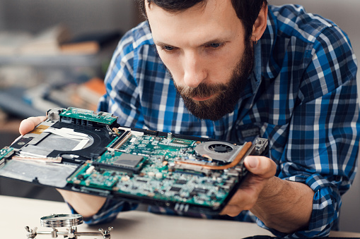 Engineer studying computer motherboard, close-up. Bearded repairman examing electronic circuit to find failure issue. Repair shop, technology, occupation concept