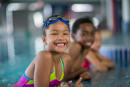 A multi-ethnic group of elementary age children are taking swimming lessons at a public pool. One girl is smiling and looking at the camera.