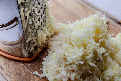 Grated potato with metal standing grater on wooden cutting board from high angle closeup