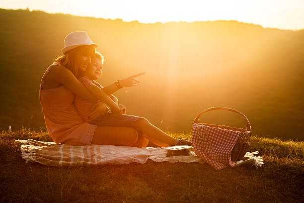 Little boy sitting on mother's lap and pointing at something while enjoying on picnic at sunset.