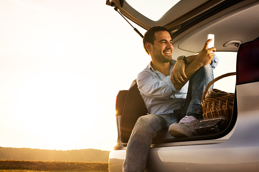 Young smiling man sitting in car trunk and reading text message on cell phone at sunset.