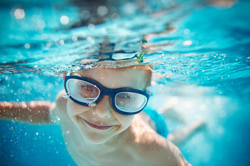 Portrait of smiling little boy enjoying underwater swim in the pool towards the camera. Sunny summer day. Copy space above the boy's head.