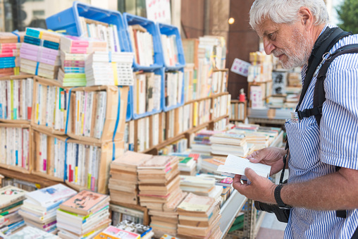 A mature man in Japan in bookstore reading a book. He has camera around his neck, lots of books on shelfs around.  