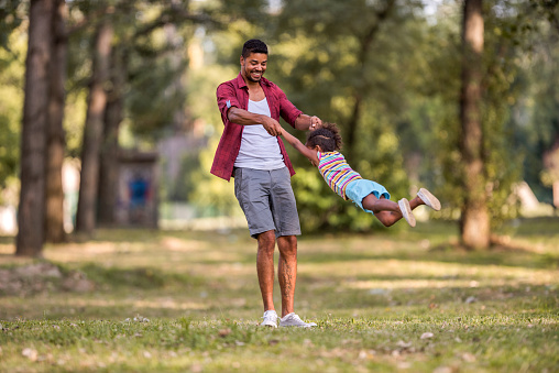 Happy African American father having fun with his small girl in nature while spinning her.