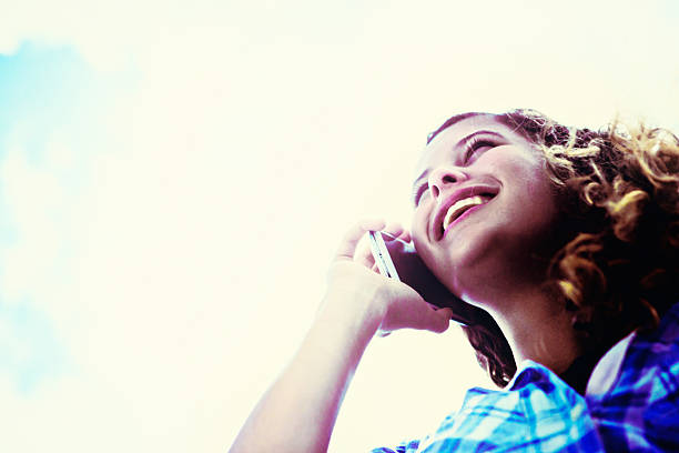 This beautiful young honey-blonde woman smiles as she chats animatedly on her smartphone, seen from directly below, against a bright background of blue sky with clouds. Copy space on the clouds.