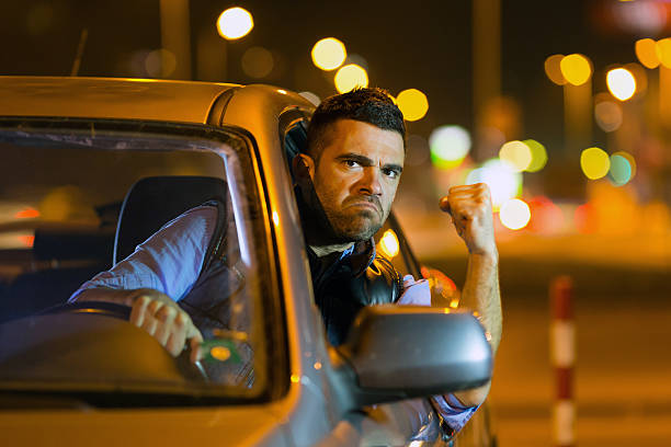 Young man driving car at night angry to traffic Man driving car at night angry to traffic, pointing at his head. Passing his head through the window. Wears casual clothes. Streets lights on background. car city urban scene commuter stock pictures, royalty-free photos & images