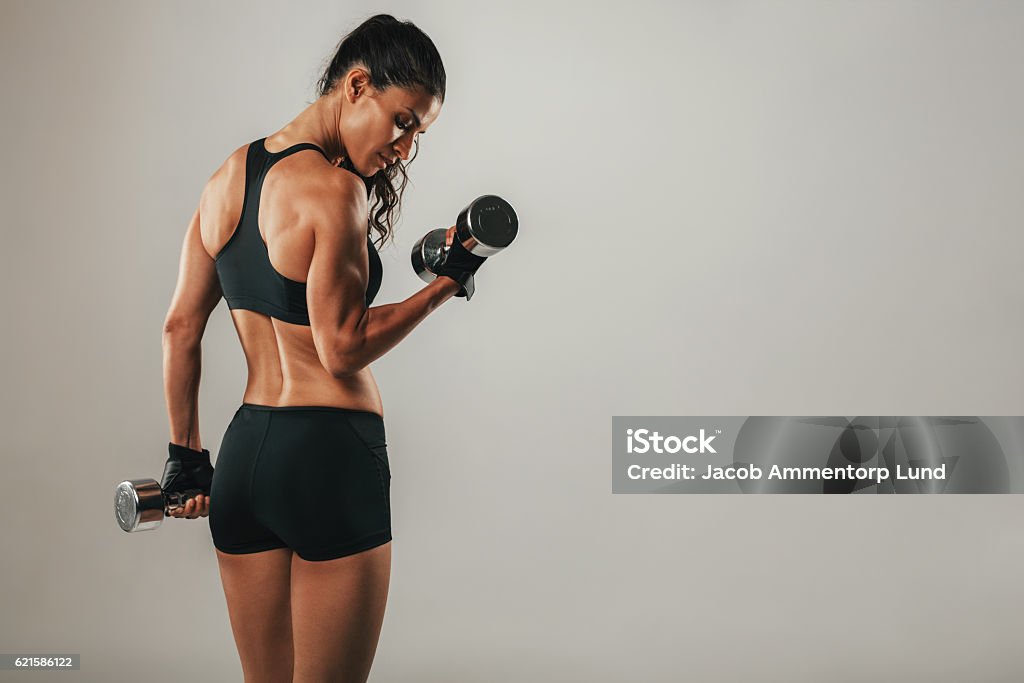 Fit strong young woman lifting weights Fit strong young woman lifting weights working out with dumbbells standing with her back to the camera flexing her arm on grey with copy space Women Stock Photo